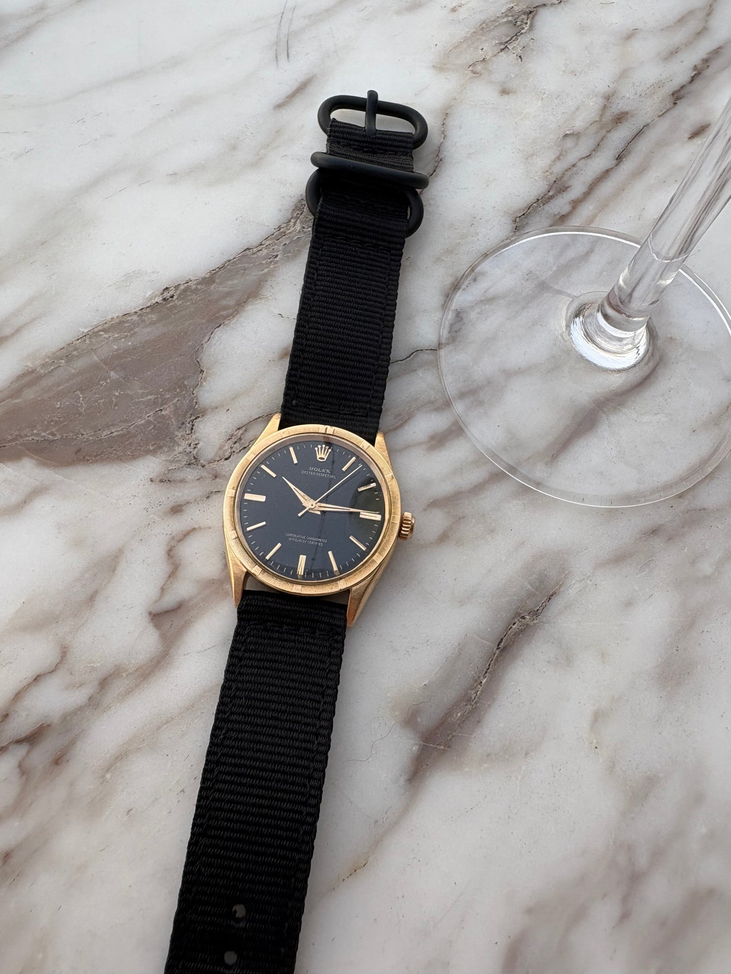 Gold watch with black strap on a marble surface next to a glass.