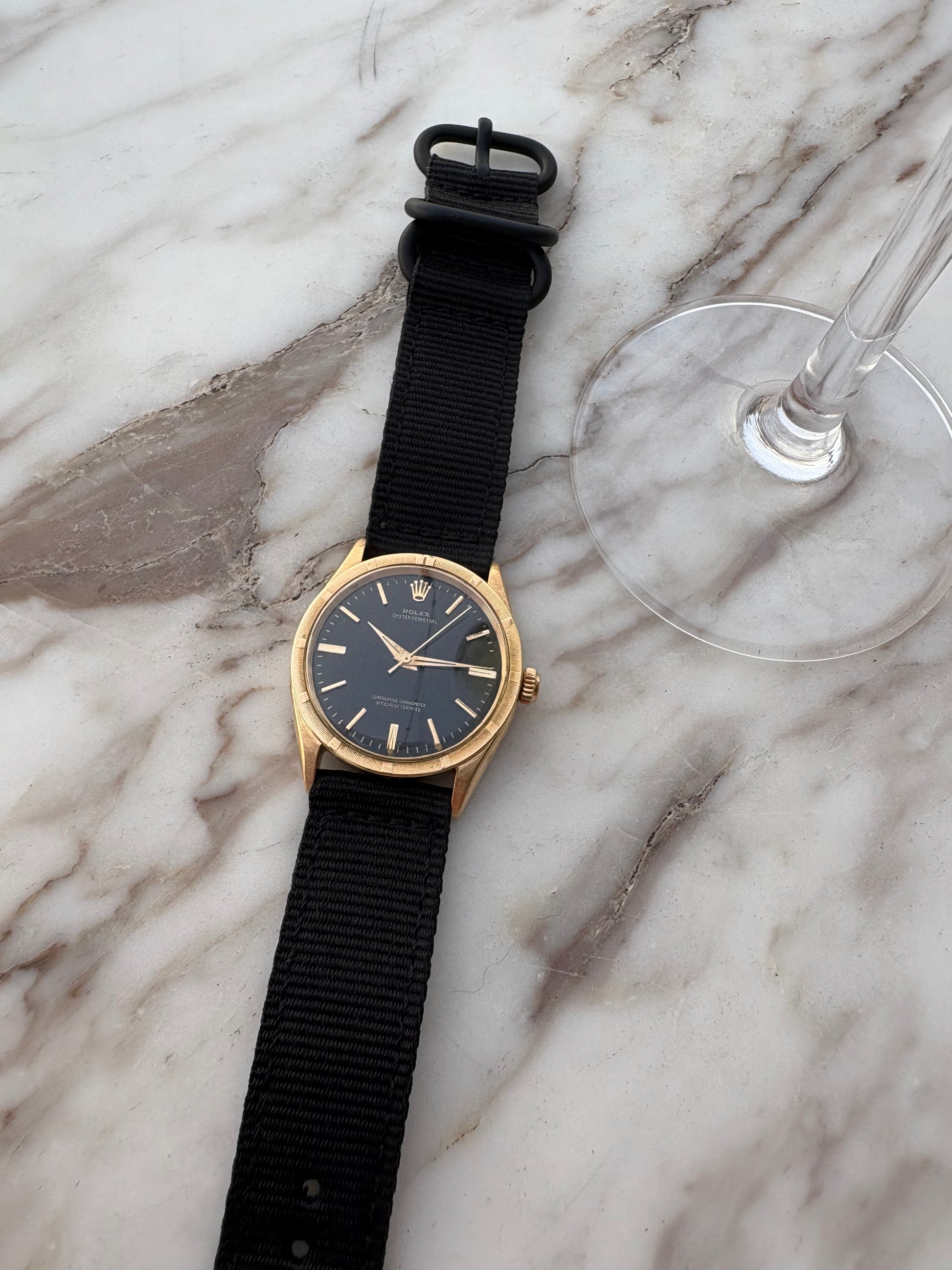 Gold watch with black strap on a marble surface next to a glass.