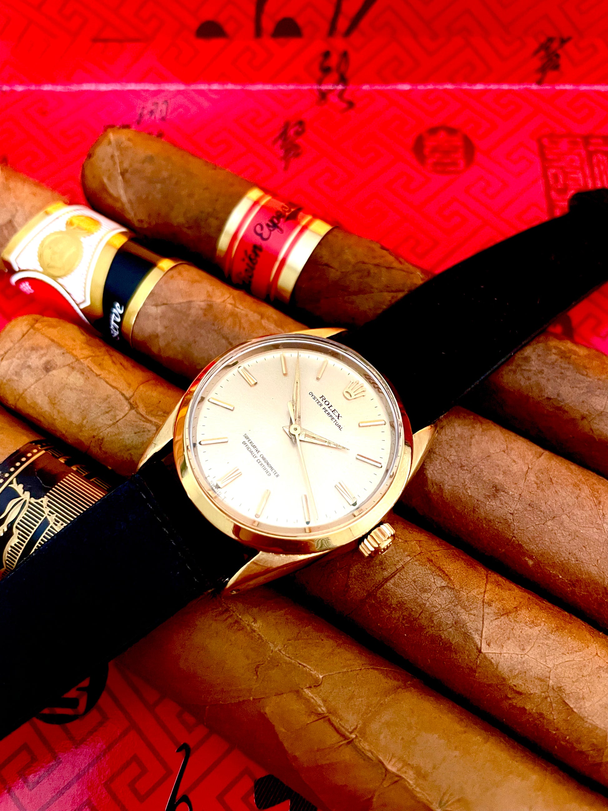 Swiss made mechanical vintage gold Rolex watch with gold bezel, gold dial, and black strap on top of cigars with a red box in the background