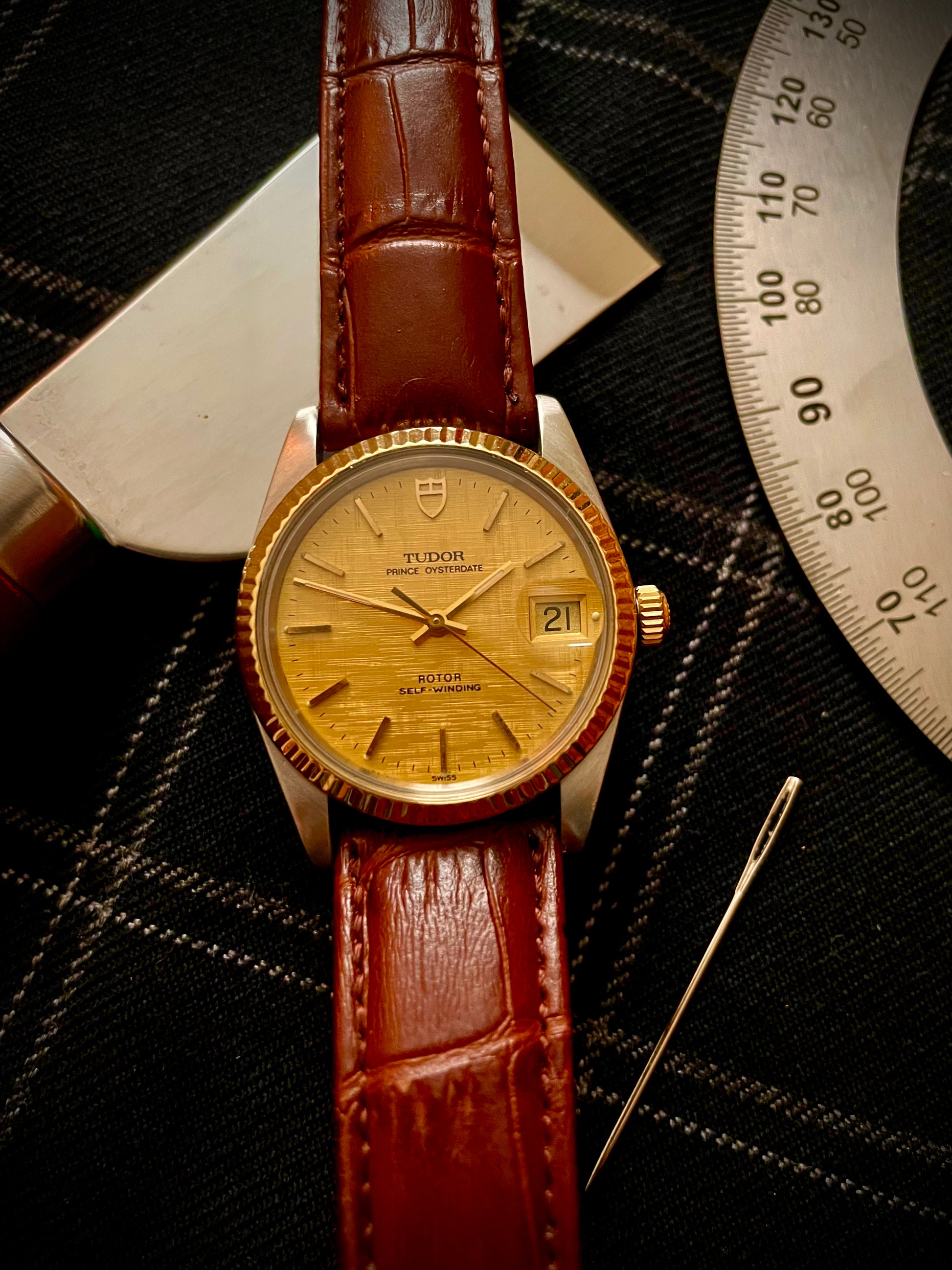 Swiss made mechanical vintage Tudor watch with gold bezel, champagne dial, and brown leather strap on a dark textured background